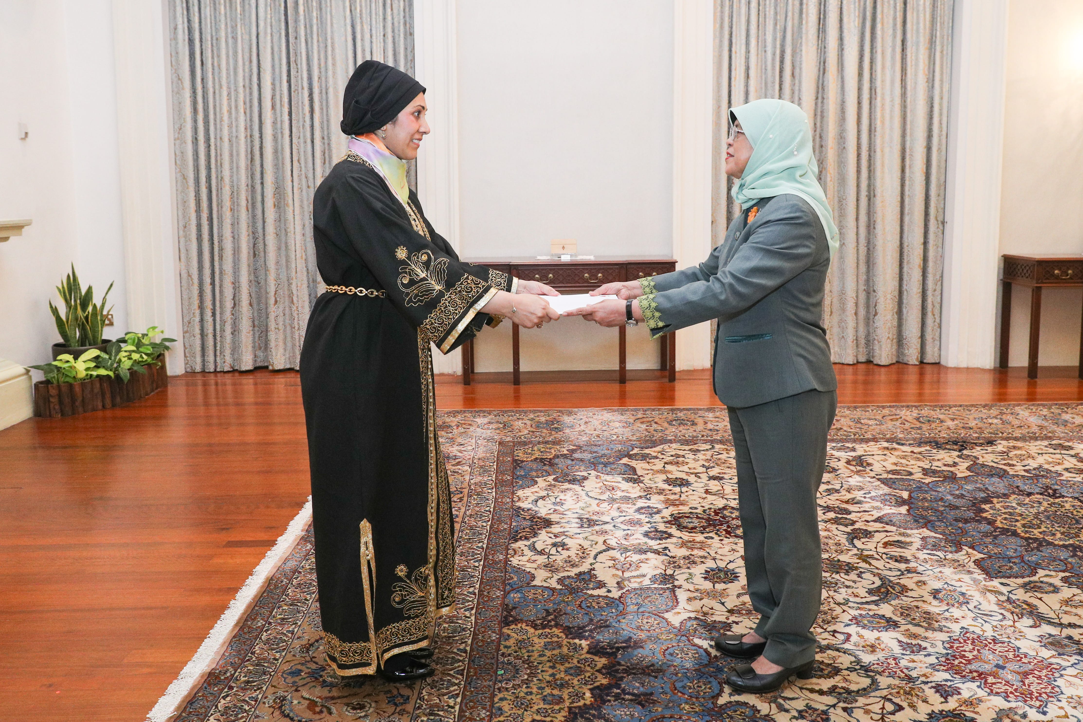 Two women exchanging document indoors, patterned rug, one wearing black and gold robe, one grey suit.